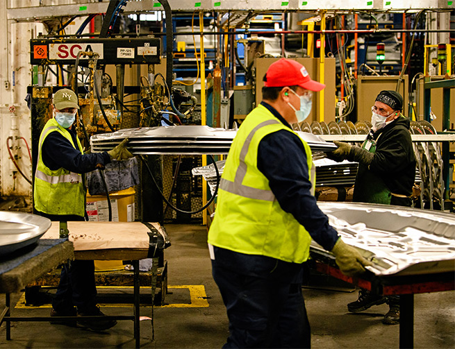 In the foreground a worker with a red hat, ear plugs, safety vest and mask walks across the photo. In the background two workers in PPE carry four sheets of silver metal