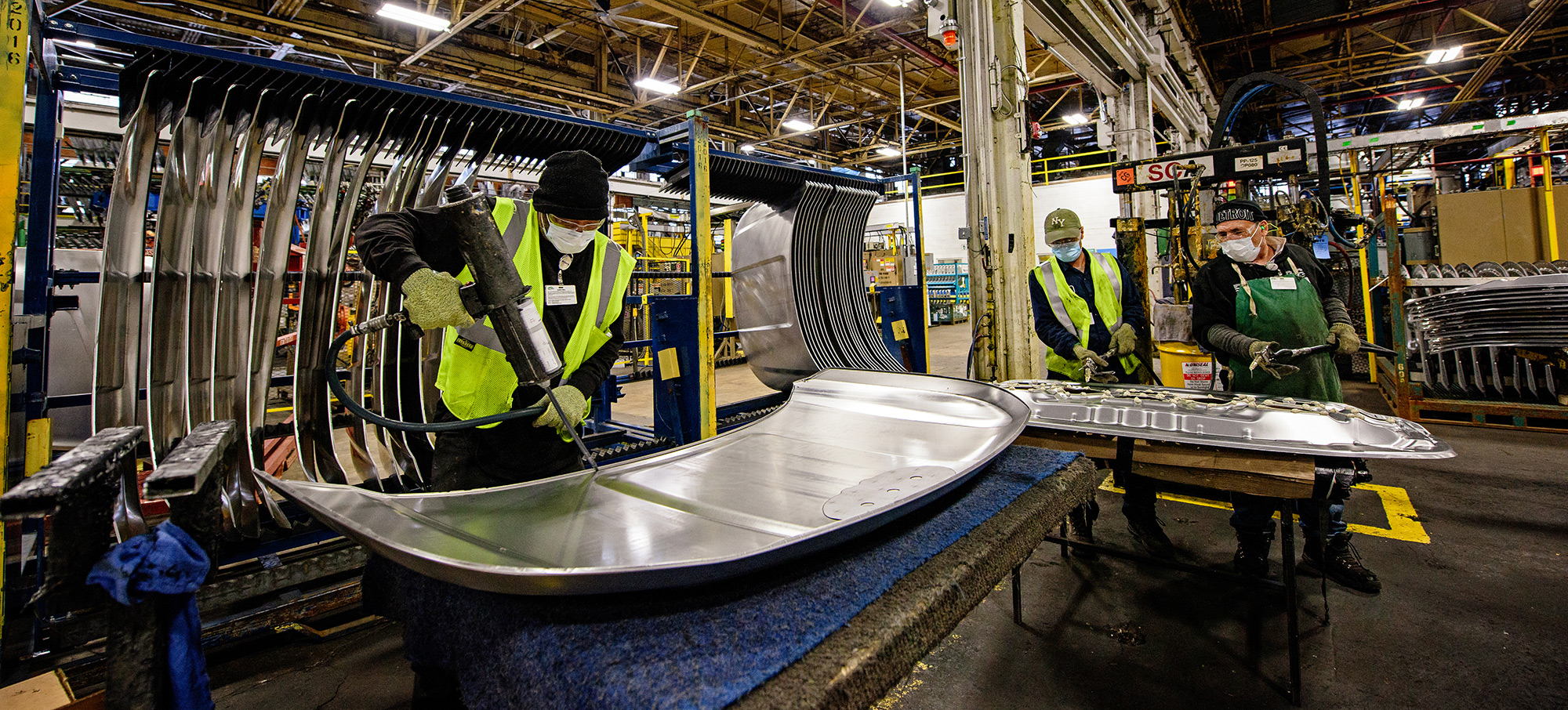 Fishbowl lens shot of three workers. One on the left in PPE has a drilling-type machine with curved silver metal on the table in front of them. Two other workers in the background work similarly on a silver sheet of metal.