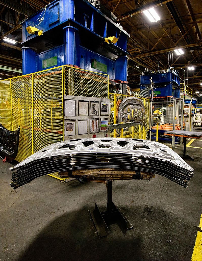 Stacks of silver curved sheet metal in front of a yellow cage holding a big blue machine.