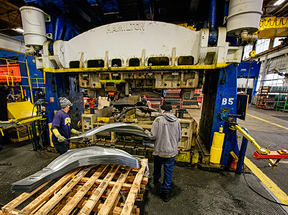 Two workers in front of a larger Hamilton stamping machine load a curved sheet of silver metal into the machine