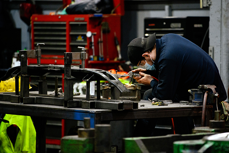 Workers wearing PPE and a baseball Carhartt cap hunches over a work table and uses a small piece of equipment for die repair Workers wearing PPE and a baseball Carhartt cap hunches over a work table and uses a small piece of equipment for die repair