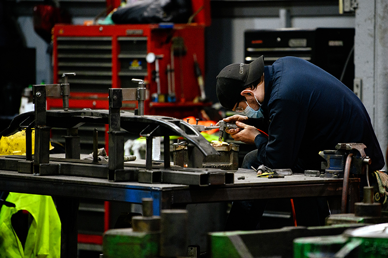 Workers wearing PPE and a baseball Carhartt cap hunches over a work table and uses a small piece of equipment for die repair