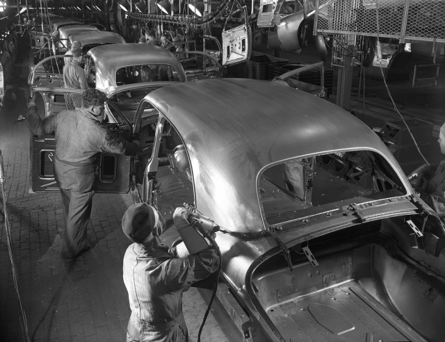 Black and white historic photo of workers using drills and inspecting vehicles in a production line