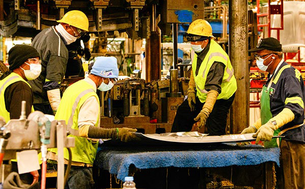 stamping-machine-02c Five men in PPE around a silver sheet of metal. Two men hold it while the three other men survey it.