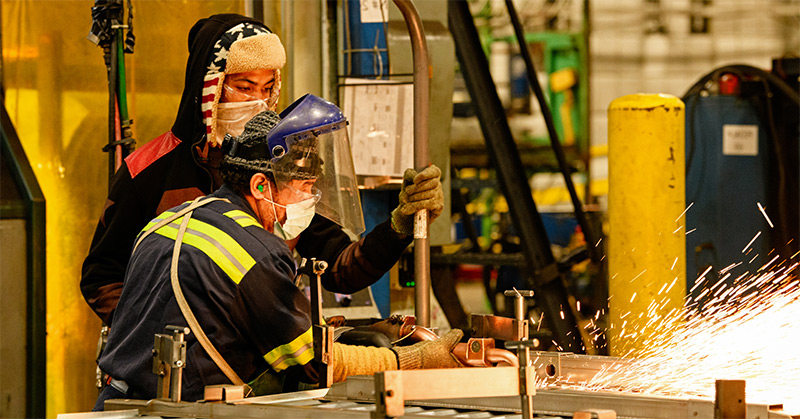 Two workers in PPE including face masks, ear plugs, safety glasses and gloves working on a welding machine with sparks flying to the right