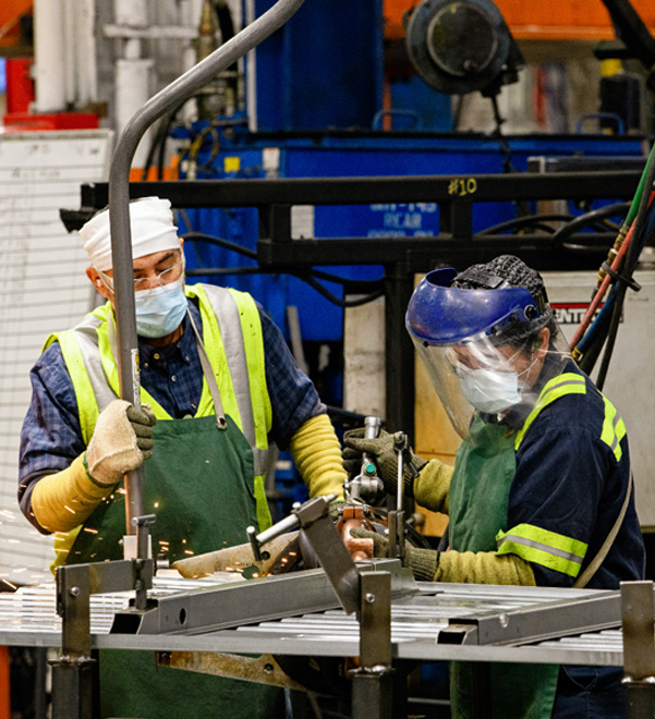 Two workers in PPE including face masks, safety glasses and aprons work with welding equipment.